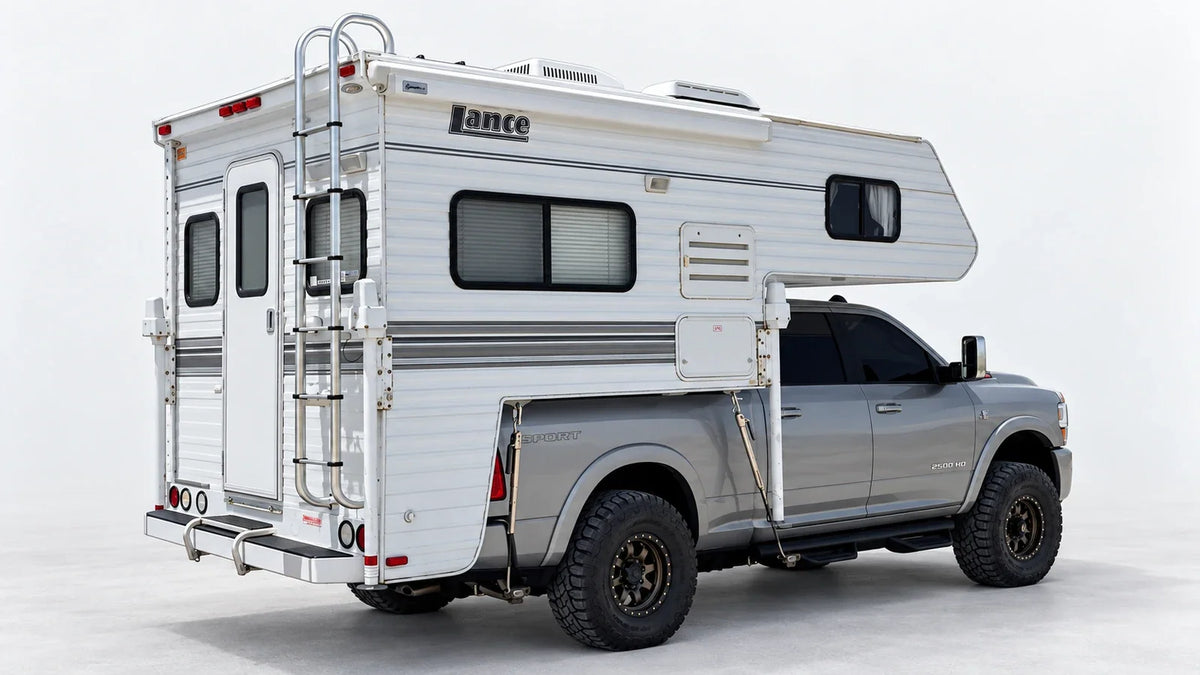 Silver pickup carrying a white slide-in camper, with front and rear turnbuckle connections visible on the near side.