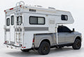 Silver pickup carrying a white slide-in camper, with front and rear turnbuckle connections visible on the near side.